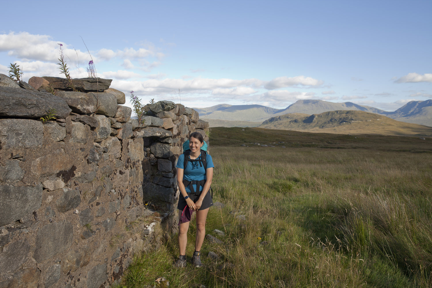 Christina stands with her hiking backpack on near the ruins of a stone building in the Scottish Highlands, with rolling grassy hills behind her.